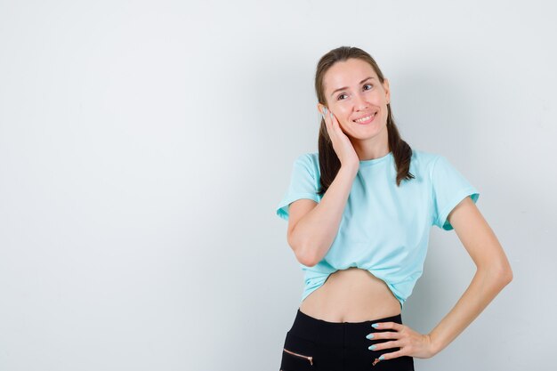 Mujer hermosa joven en camiseta con la mano en la cara, mirando a otro lado y mirando alegre, vista frontal.