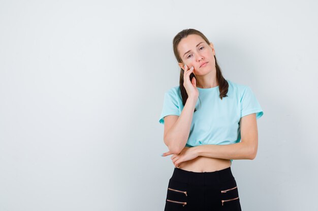 Mujer hermosa joven en camiseta, pantalones con los dedos en la cara y mirando triste, vista frontal.