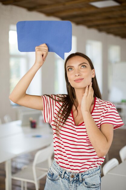 Mujer hermosa joven en camiseta a rayas mirando soñando hacia arriba sosteniendo un mensaje en forma de papel azul en la mano debajo de la cabeza en el trabajo en una oficina vacía moderna