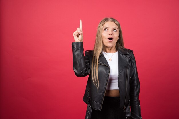 Mujer hermosa joven con chaqueta de cuero de pie sobre la pared roja y apuntando con el dedo hacia arriba