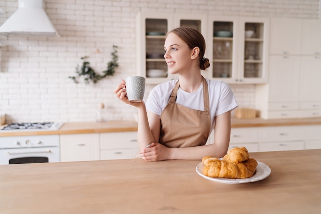Mujer hermosa joven en la cocina en un delantal con café y croissant disfruta de su mañana