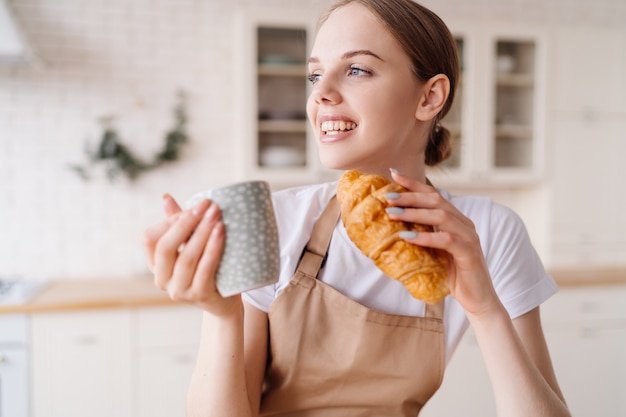 Mujer hermosa joven en la cocina en un delantal con café y croissant disfruta de su mañana