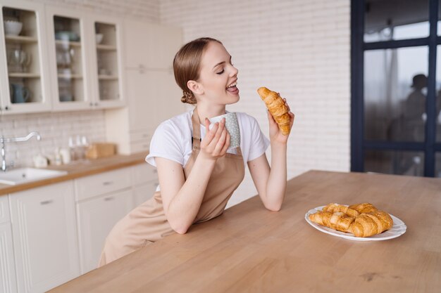 Mujer hermosa joven en la cocina en un delantal con café y croissant disfruta de su mañana