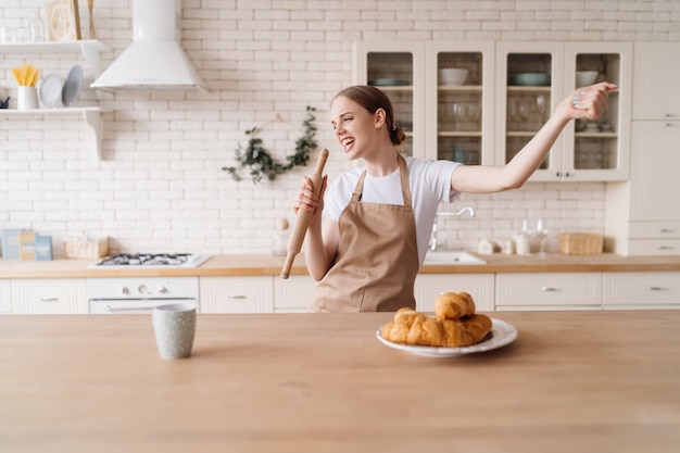 Foto gratuita mujer hermosa joven en la cocina en un delantal canta en un rodillo de masa