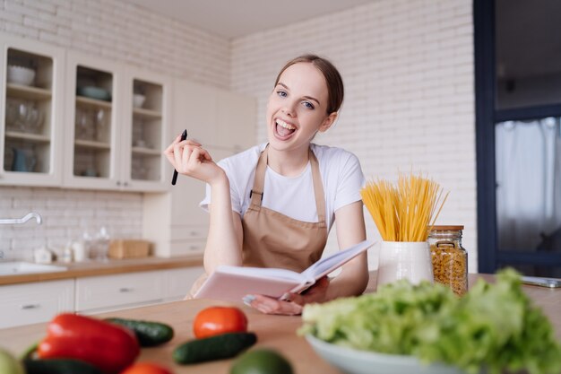 Mujer hermosa joven en la cocina en un delantal escribe sus recetas favoritas junto a verduras frescas