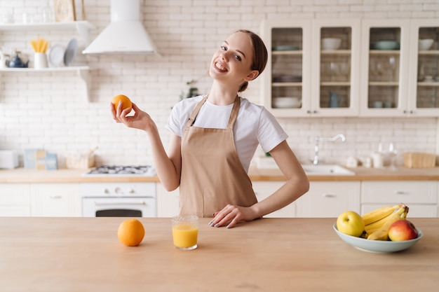 Mujer hermosa joven en la cocina en un delantal, frutas y jugo de naranja