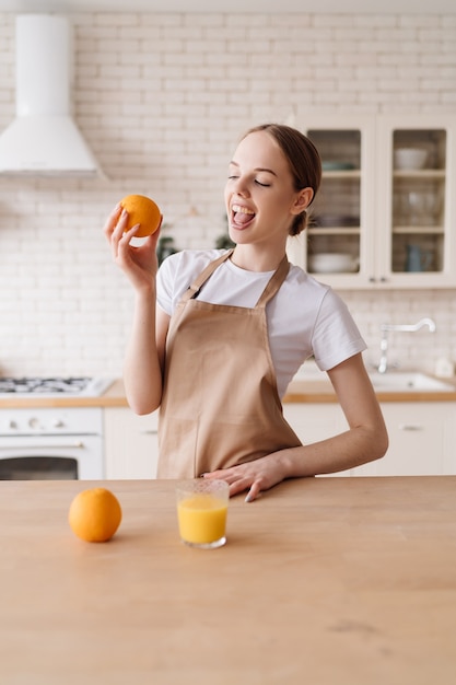 Mujer hermosa joven en la cocina en un delantal, frutas y jugo de naranja