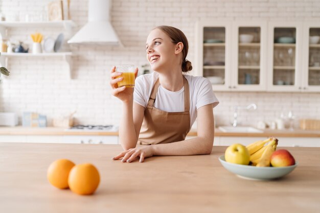 Mujer hermosa joven en la cocina en un delantal, frutas y jugo de naranja