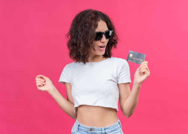 Mujer hermosa joven complacida con gafas de sol y sosteniendo la tarjeta en la pared rosa aislada