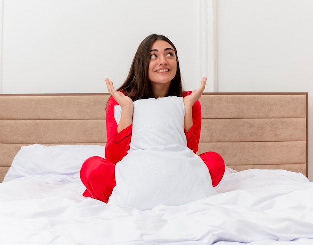 Foto gratuita mujer hermosa joven complacida en pijama rojo sentado en la cama con almohada mirando hacia arriba sonriendo con cara feliz disfrutando de fin de semana en el interior del dormitorio