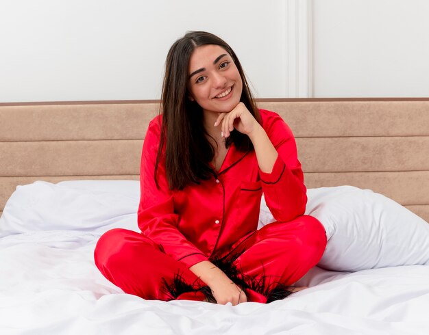 Mujer hermosa joven complacida en pijama rojo sentado en la cama mirando a la cámara sonriendo con cara feliz en el interior del dormitorio sobre fondo claro