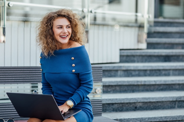 Mujer hermosa joven con una computadora portátil se sienta en un banco en la parte comercial de la ciudad. Mujer hermosa joven, autónomo, trabaja en la computadora portátil en verano usar vestido azul. Concepto de estilo de vida.