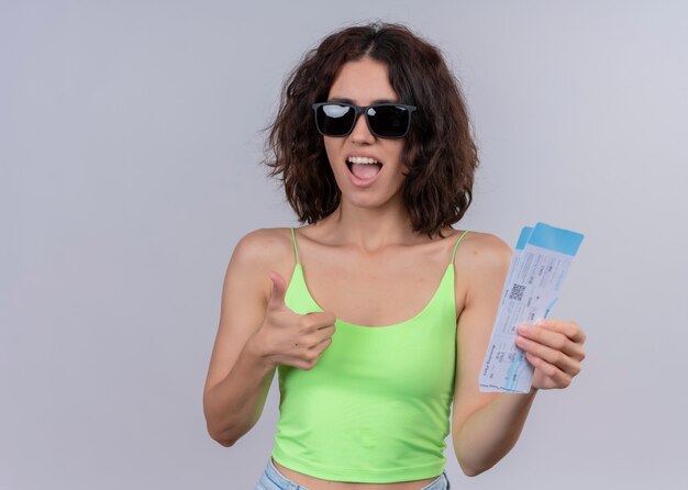 Mujer hermosa joven confidente que lleva gafas de sol y que sostiene los billetes de avión y que muestra el pulgar hacia arriba en la pared blanca aislada con el espacio de la copia