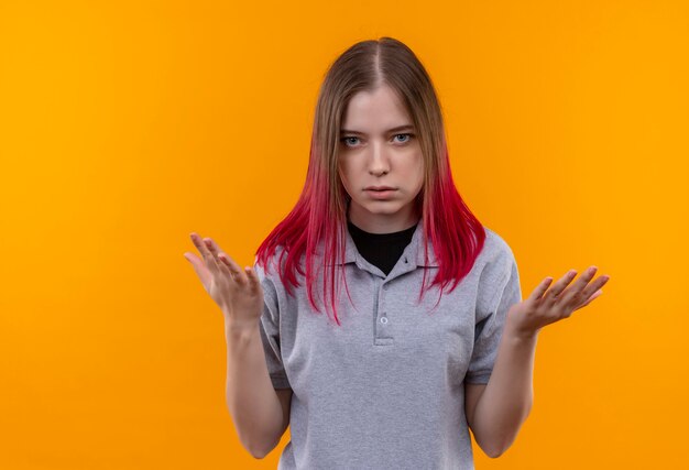 Mujer hermosa joven confundida que lleva la camiseta gris extiende las manos en la pared amarilla aislada