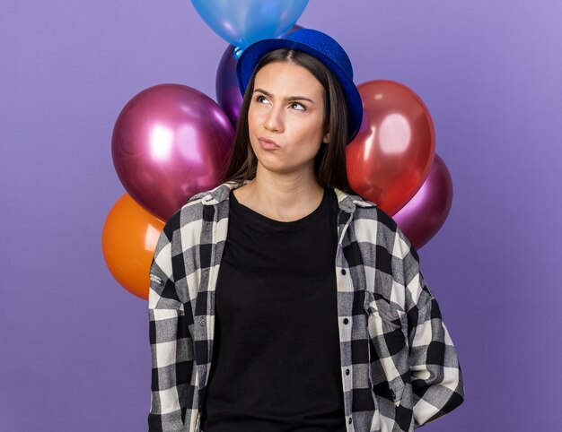 Mujer hermosa joven confundida que lleva el sombrero del partido que se coloca delante de los globos aislados en la pared azul