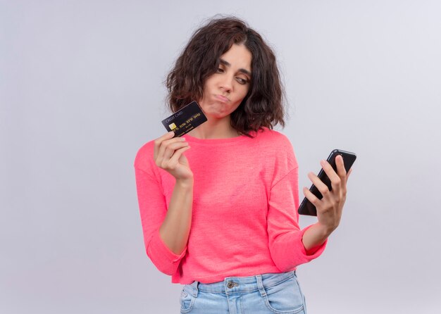 Mujer hermosa joven confundida que sostiene la tarjeta y el teléfono móvil en la pared blanca aislada con el espacio de la copia