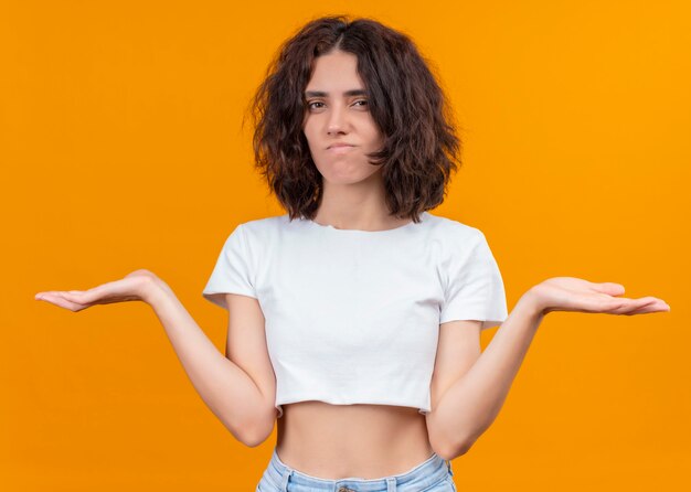 Mujer hermosa joven confusa que muestra las manos vacías en la pared naranja aislada