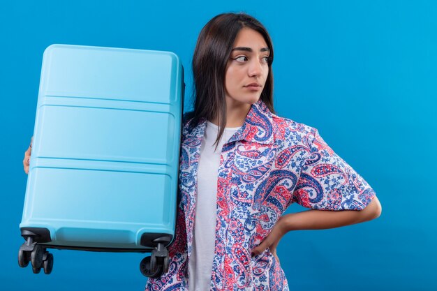 Mujer hermosa joven confusa que sostiene la maleta del viaje que mira a un lado con la expresión facial dudosa que se coloca sobre el espacio azul aislado