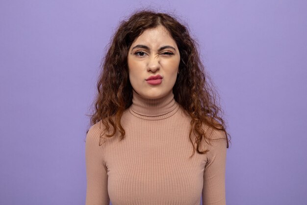 Mujer hermosa joven en cuello alto beige haciendo boca torcida con expresión decepcionada de pie en púrpura