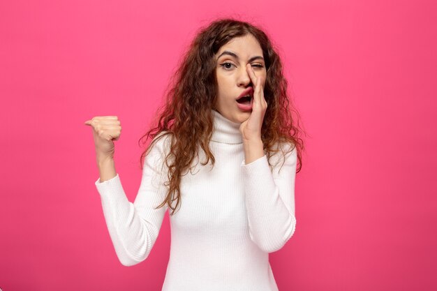 Mujer hermosa joven en cuello alto blanco contando un secreto sosteniendo la mano en la boca apuntando con el pulgar hacia el lado de pie en rosa