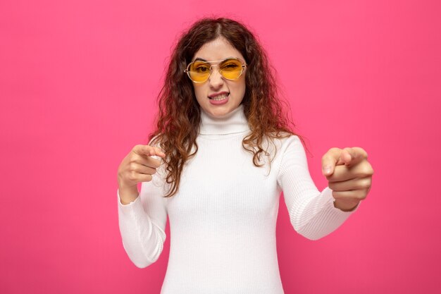 Mujer hermosa joven en cuello alto blanco con gafas amarillas mirando apuntando con los dedos índices al frente con expresión molesta