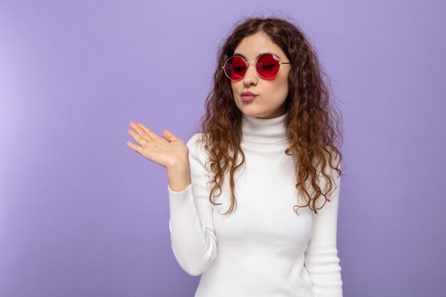 Mujer hermosa joven en cuello alto blanco con gafas rojas mirando su brazo presentando algo que parece seguro