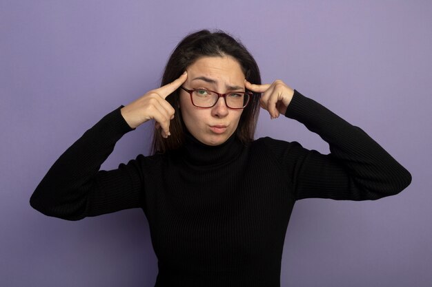 Mujer hermosa joven en un cuello alto negro y gafas apuntando a sus sienes mirando al frente con expresión escéptica de pie sobre la pared púrpura