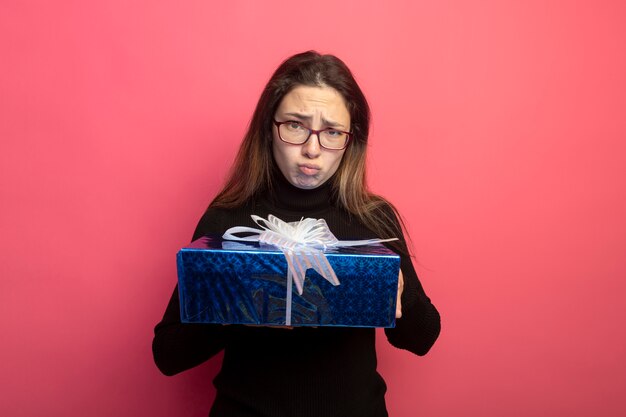 Mujer hermosa joven en un cuello alto negro y gafas con caja de regalo mirando al frente con expresión triste estar disgustado de pie sobre la pared rosa