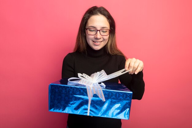 Mujer hermosa joven en un cuello alto negro y gafas con caja de regalo mirándola con cara feliz de pie sobre la pared rosa