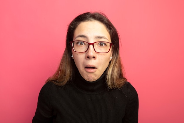 Mujer hermosa joven en un cuello alto negro y gafas mirando al frente confundido parado sobre pared rosa