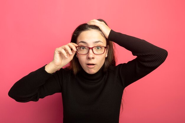 Mujer hermosa joven en un cuello alto negro y gafas mirando al frente confundido tocando sus gafas de pie sobre la pared rosa