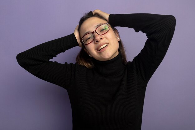 Mujer hermosa joven en un cuello alto negro y gafas mirando al frente feliz y positivo con las manos en la cabeza sonriendo de pie sobre la pared púrpura