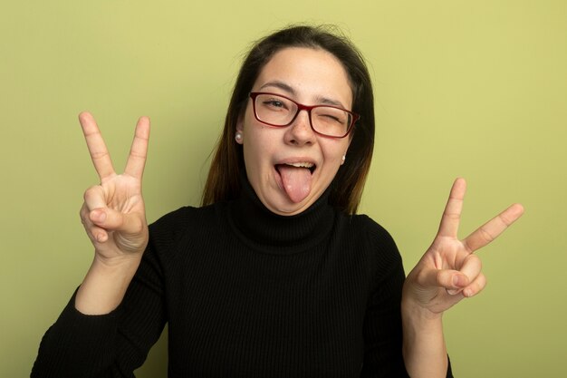 Mujer hermosa joven en un cuello alto negro y gafas mirando al frente feliz y positivo mostrando el signo v sacando la lengua de pie sobre la pared de luz