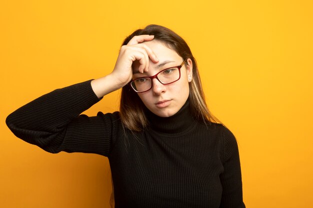 Mujer hermosa joven en un cuello alto negro y gafas mirando a un lado desconcertado con la mano en la cabeza de pie sobre la pared naranja