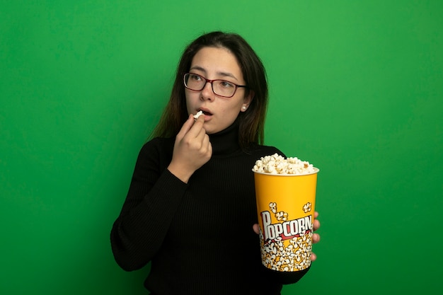 Mujer hermosa joven en un cuello alto negro y gafas sosteniendo un balde con palomitas de maíz comiendo palomitas de maíz mirando a un lado con feliz y posiitve de pie sobre la pared verde