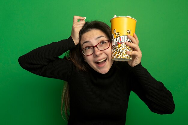 Mujer hermosa joven en un cuello alto negro y gafas sosteniendo un balde con palomitas de maíz mirando hacia arriba feliz y emocionado de pie sobre la pared verde