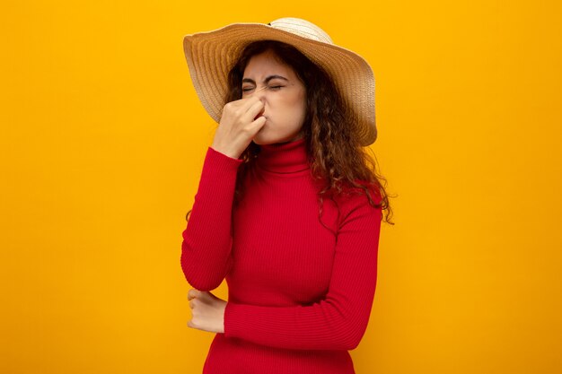 Mujer hermosa joven en cuello alto rojo con sombrero de verano cerrando la nariz con los dedos que sufren de hedor