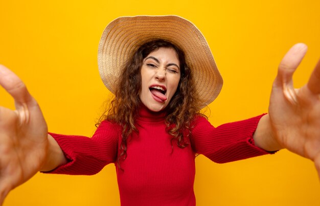 Mujer hermosa joven en cuello alto rojo con sombrero de verano feliz y alegre divirtiéndose sacando la lengua de pie en naranja