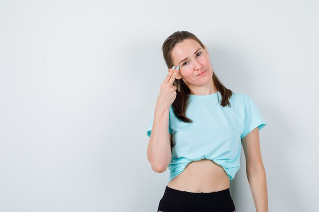 Mujer hermosa joven con los dedos en las sienes en camiseta y mirando contento. vista frontal.