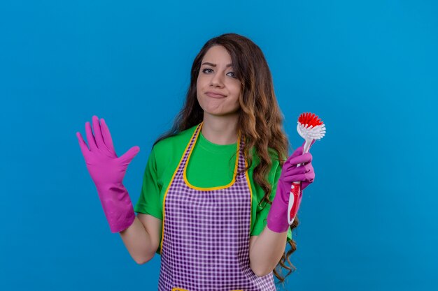 Mujer hermosa joven disgustada con pelo largo ondulado en delantal y guantes sosteniendo cepillo de fregar mirando confundido de pie con las manos levantadas de pie en azul