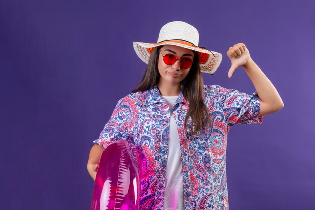 Mujer hermosa joven disgustada con sombrero de verano y gafas de sol rojas con anillo inflable mostrando los pulgares hacia abajo sobre la pared púrpura