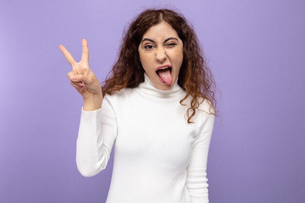 Mujer hermosa joven divertida en cuello alto blanco que muestra el signo v sacando la lengua de pie en púrpura