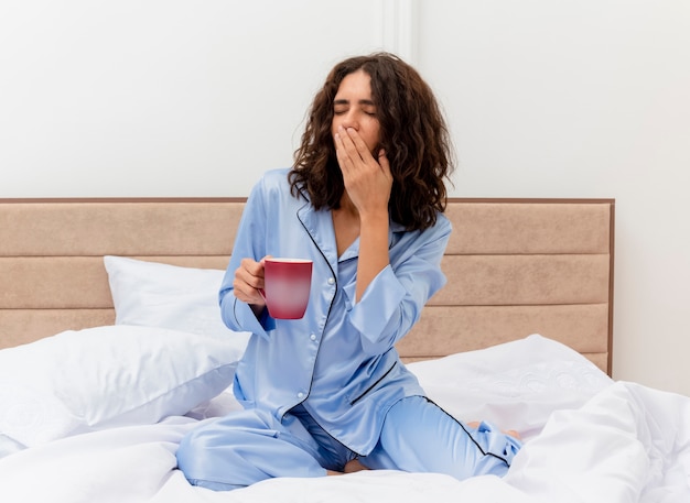 Mujer hermosa joven divertida en pijama azul sentada en la cama con una taza de café despertando bostezando sensación de fatiga matutina en el interior del dormitorio