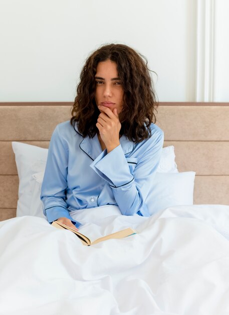 Mujer hermosa joven divertida en pijama azul sentado en la cama con el libro mirando a la cámara con cara seria en el interior del dormitorio sobre fondo claro