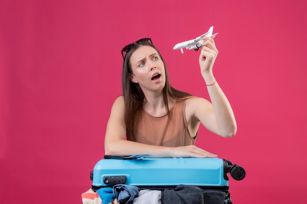 Mujer hermosa joven dudosa con maleta de viaje llena de ropa con avión de juguete mirando incierto sobre espacio rosa