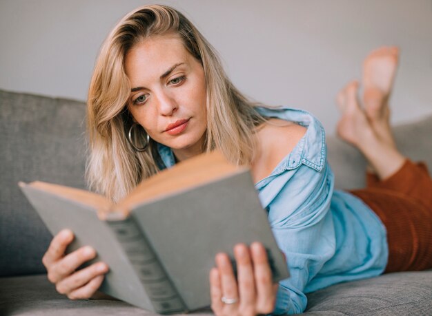 Mujer hermosa joven elegante que miente en el sofá que lee el libro