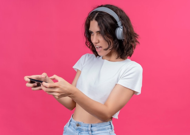 Mujer hermosa joven emocionada que lleva puestos los auriculares y que sostiene el teléfono móvil que mira el lado izquierdo en la pared rosada