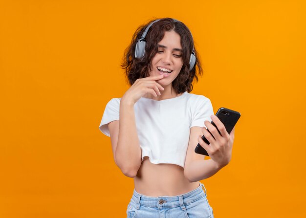 Mujer hermosa joven emocionada que lleva puestos los auriculares que sostienen el teléfono móvil y que pone el dedo en el labio en la pared anaranjada aislada con el espacio de la copia