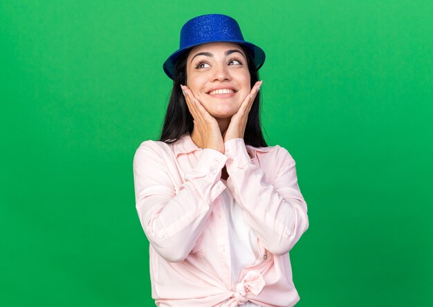 Mujer hermosa joven emocionada que lleva el sombrero del partido que pone las manos en las mejillas aisladas en la pared verde