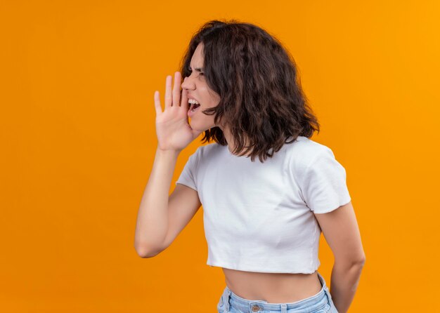 Mujer hermosa joven enojada gritando en el lado izquierdo con la mano cerca de la boca en la pared naranja aislada con espacio de copia
